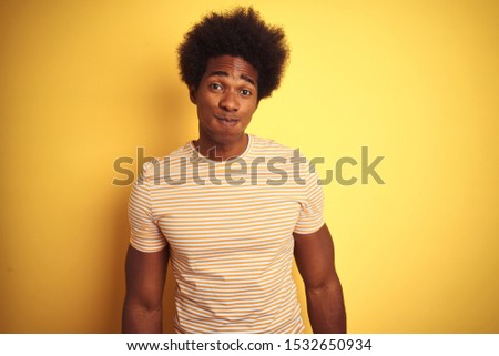 American man with afro hair wearing striped t-shirt standing over isolated yellow background puffing cheeks with funny face. Mouth inflated with air, crazy expression.