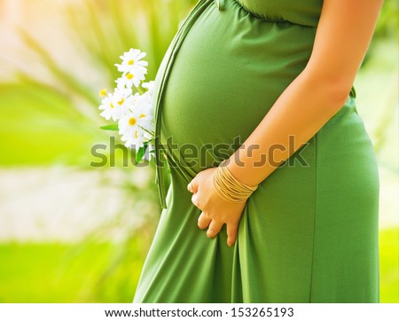 Closeup on tummy of pregnant woman, wearing long green dress, holding in hands bouquet of daisy flowers outdoors, new life concept Royalty-Free Stock Photo #153265193