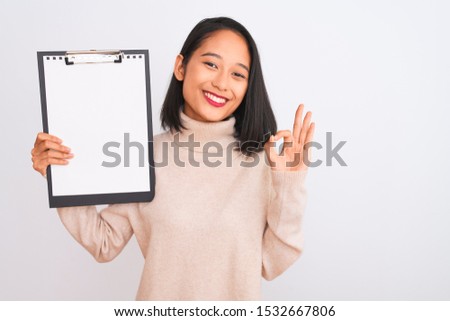 Young chinese inspector woman holding clipboard standing over isolated white background doing ok sign with fingers, excellent symbol