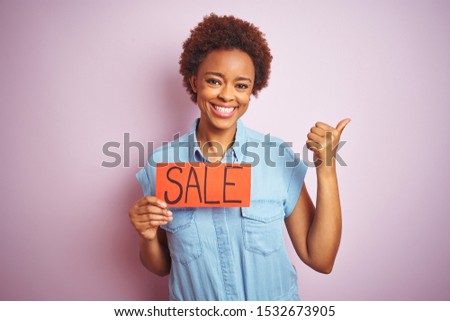 African american woman holding sale board over pink isolated background pointing and showing with thumb up to the side with happy face smiling