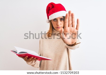 Beautiful redhead woman wearing christmas hat and reading a book over isolated background with open hand doing stop sign with serious and confident expression, defense gesture