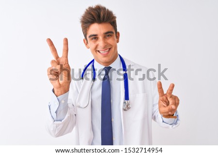 Young handsome doctor man wearing stethoscope over isolated white background smiling looking to the camera showing fingers doing victory sign. Number two.