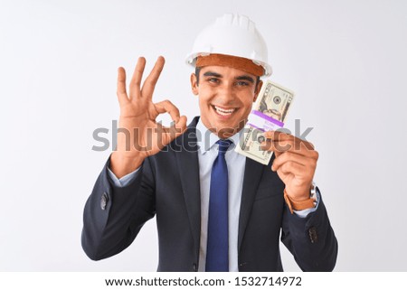 Young handsome architect man wearing helmet holding dollars over isolated white background doing ok sign with fingers, excellent symbol