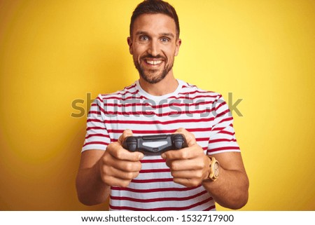 Young handsome man playing video games using joystick game pad over yellow background with a happy face standing and smiling with a confident smile showing teeth