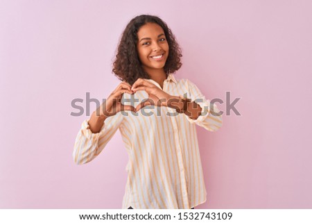 Young brazilian woman wearing striped shirt standing over isolated pink background smiling in love doing heart symbol shape with hands. Romantic concept.