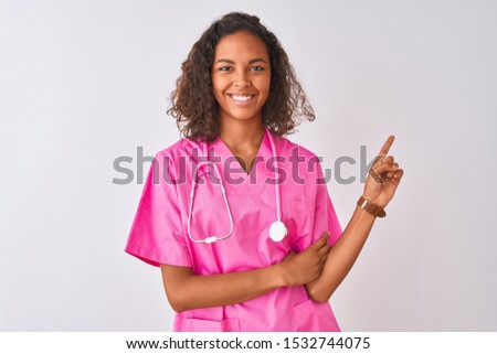 Young brazilian nurse woman wearing stethoscope standing over isolated white background with a big smile on face, pointing with hand and finger to the side looking at the camera.