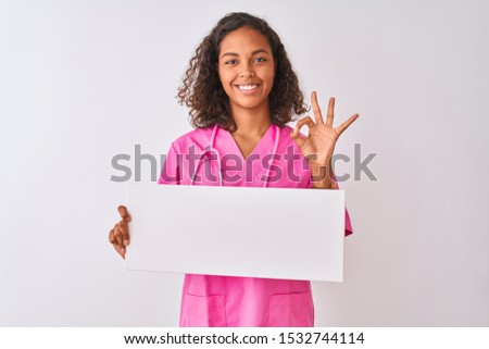 Young brazilian nurse woman holding banner standing over isolated white background doing ok sign with fingers, excellent symbol
