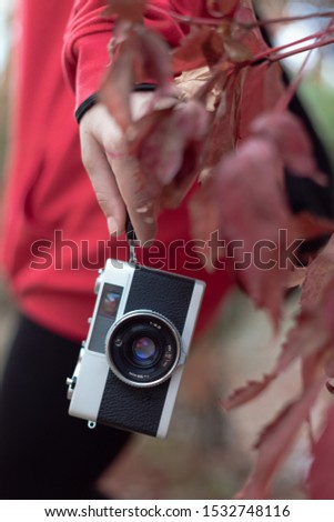 Telemetric film camera with strap hanging from the hand of a girl dressed in red. Autumnal leaves also appear on the scene.