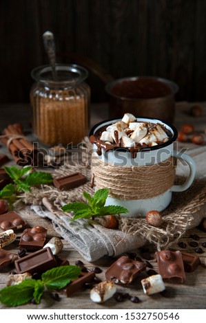 On the table, on a napkin, is a mug of hot chocolate and marshmallows. Around chocolate, coffee, marshmallows, nuts and mint petals. Behind a glass jar with sugar and a spoon, cinnamon and a ladle.