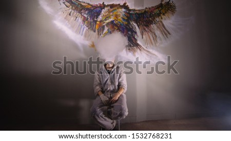 Artist designer draws an eagle on the wall. Craftsman decorator paints a picture with acrylic oil color. Vaper smokes vape. Painter painter dressed in a paint coat. Indoor. Dark magic cinematic look.