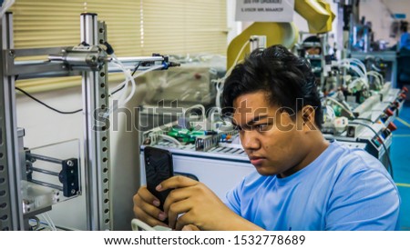 A young Malay engineering student working using a smartphone taking photo of automation machine system at the laboratory. Image contain noise reduction and bit out of focus.