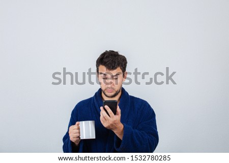 Portrait of sleepy young man messy hair, wears blue bathrobe, drinking coffee from his cup and speking on phone isolated on white background.