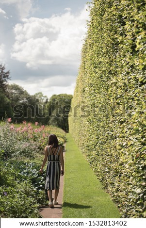Woman on a stripy summer dress walks on a path next to a tall hedge and flowers in Princes Street Gardens, Edinburgh, Scotland, UK