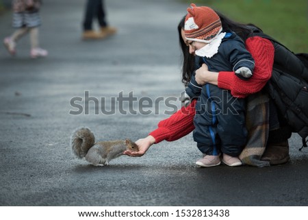 A Chinese mother Holds her baby wearing a winter jumpsuit, gloves and a fox hat as she feeds a brown squirrel which eats from her hand In Princes Street Gardens, Edinburgh, Scotland, United Kingdom