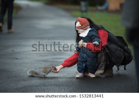 A mother and baby wearing winter clothes feed a brown squirrel In Princes Street Gardens, Edinburgh, Scotland, UK
