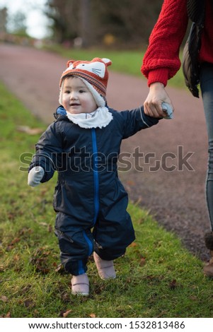 An Asian baby wearing winter clothes, a fox hat and mittens holds her mom’s hand while smiling and walking on grass in Princes Street Gardens, Edinburgh, Scotland, UK