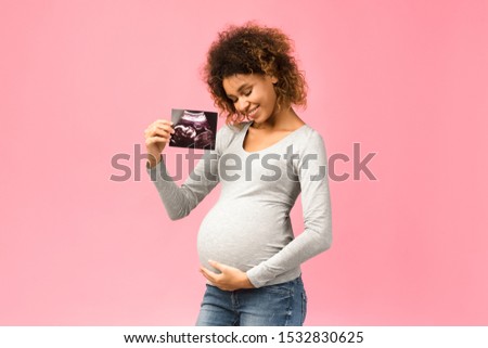 Happy afro pregnant woman with her baby sonography, enjoying first photo of her unborn child, pink background
