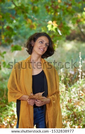 
Latin brunette woman with a black t-shirt in a forest with dry leaves and autumnal colors, the sun enters between the branches of the trees.