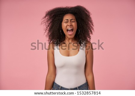 Stressed young dark skinned woman with curly hair posing over pink background with hands down, frowning her face with closed eyes and screaming loud with wide mouth opened