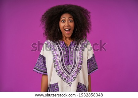 Portrait of surprised curly dark skinned female in casual clothes posing over purple background, keeping her hands along body, raising eyebrows and looking at camera with wide eyes opened