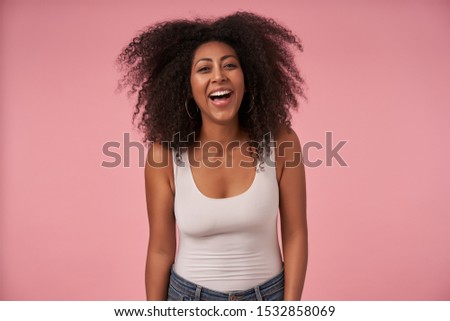 Joyful pretty young dark skinned woman with curly hair wearing casual clothes standing over pink background, keeping hands along her body and smiling happily to camera