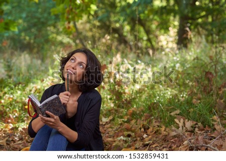 
Latin brunette woman with a black t-shirt in a forest with dry leaves and autumnal colors, the sun enters between the branches of the trees, she is sitting with a notebook in her hands.