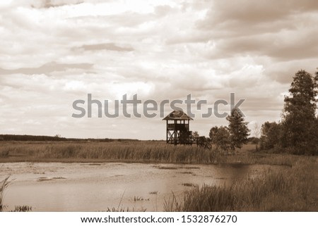 Sepia photo of a vast yet shallow river or lake with its banks covered with reeds and grass and a single observation or sentry tower seen on a cloudy, windy day on a Polish countryside