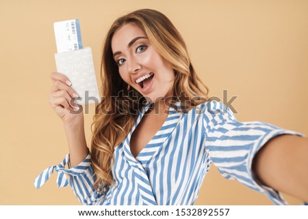 Portrait of positive woman holding passport and airline ticket while taking selfie photo isolated over beige background in studio