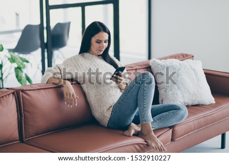Profile side photo of concentrated girl use her smartphone have inline conversation with friends write sms sit on brown leather divan in house indoors wear knitted sweater denim jeans