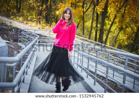 A young slender blond woman in a pink sweater with the inscription in Italian "I love you" and a black tulle skirt is standing straightening her hair against the background of autumn trees and railing