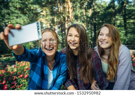Three sisters take a selfie on smartphone sitting in a park on a background of flowers on sunny day. Brunette, blonde and redhead girl smile at camera. Friendship, family walks outdoor.