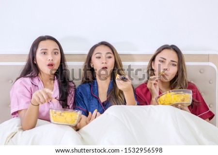 Group of beautiful teenage girls sitting on the bed while watching TV and enjoying snack in the bedroom