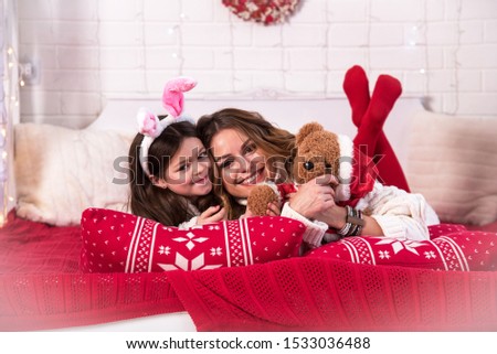 Smiling joyful mom with daughter in Christmas ears with soft toy teddy bear in bed with illuminations (New Year, 2020, Christmas concept)
