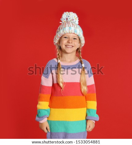 Cute little girl in hat and warm sweater on red background. Winter season