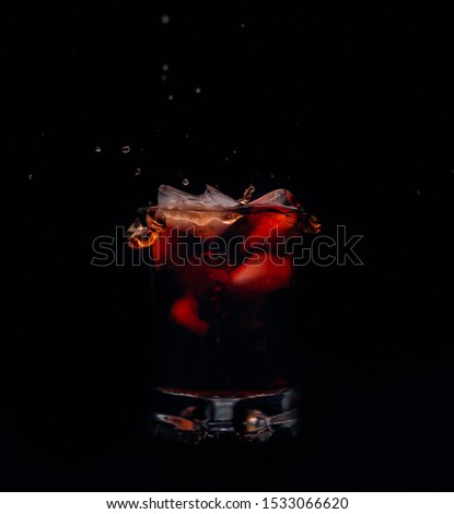 Glass of red drink with some ice falling and creating splashes and some water drops around separated on black background. 