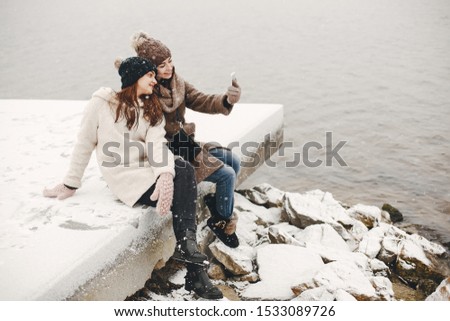 two bright and merry girls sitting in the frozen snowy park neaw water and use the phone