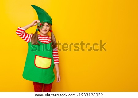 Portrait of a beautiful cheerful little girl dressed as a Christmas elf, holding a mistletoe, isolated on yellow colored background with copy space