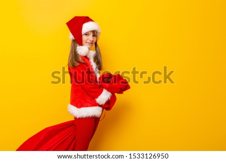 Little girl dressed as Santa carrying a huge sack of Christmas presents isolated on yellow colored background