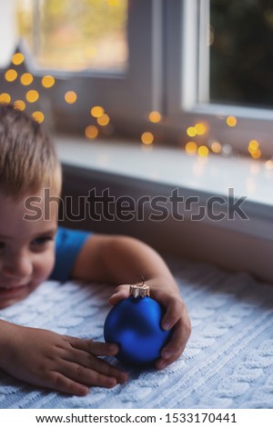 Happy child boy holding blue Christmas ball near window indoor with warm garland lights on blurred background. Participation of children in preparation of home holiday. Cozy New Year atmosphere.