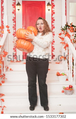 adult cute fat girl in a white sweater with gold stars holds a stack of large pumpkins and stands on the porch of her white house.