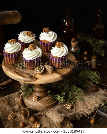homemade carrot or pumpkin cupcakes with white cream and walnut on top in purple cupcake holders on wooden cake stand on rustic table with juniper branches, nuts, old bottles, spoons