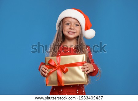 Happy little child in Santa hat with gift box on blue background. Christmas celebration