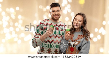christmas, people and holidays concept - portrait of happy couple at ugly sweater party with cupcakes over festive lights background