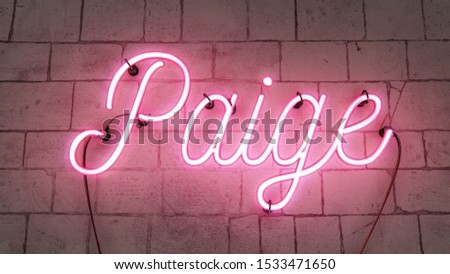 Bright pink neon sign spelling the girls name of Paige, on a concrete brick grunge background.