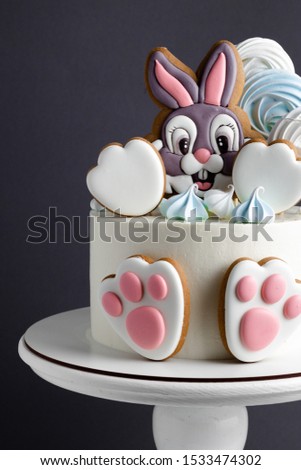 White cake with bunny gingerbread cookie on top and bunny paw cookies on the front. Dark grey background behind.