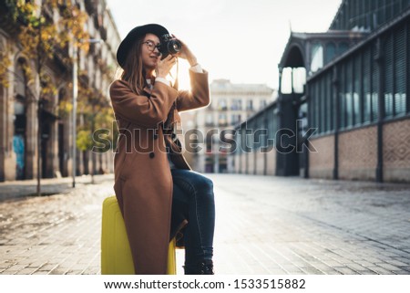 Photographer tourist with suitcase take photo on retro camera. Smiling girl in hat travels in Barcelona. Sunlight flare street in europe city. Traveler hipster shooting architecture, copy space mockup