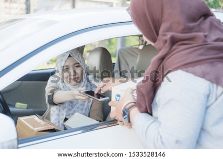 Thai young muslim custom wearing hijab female signing signature on smart phone to receive package from delivery female worker from the car.