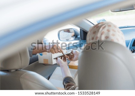 Thai young muslim custom wearing hijab female signing signature on smart phone to receive package from delivery female worker from the car.