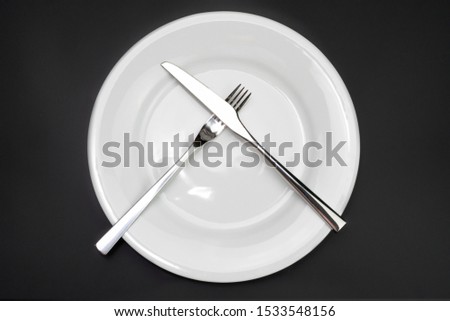 Table setting. Empty plate, knife and fork on a black background. The fork and knife lie crosswise on a plate, the meal is finished, the dish was not pleasant. Top view and flat lay with copy space.