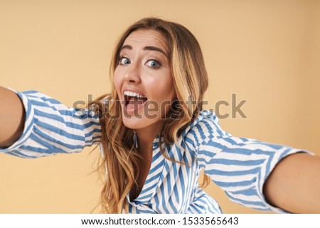 Portrait of an attractive excited young woman with long curly blonde hair wearing casual clothing standing isolated over beige background, taking a selfie
